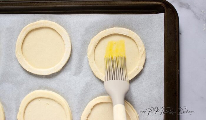 Place whole rounds on a parchment-lined baking sheet. Brush lightly with beaten egg. Place a ring on top of each base and brush the top ring with egg wash too (avoid brushing the sides to allow full puffing). Prick the center (not the ring) lightly with a fork to prevent too much puffing inside.