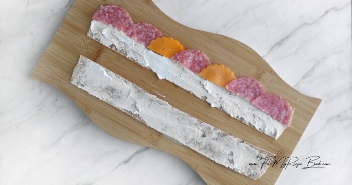 Place the tortilla on the board and trim off the edges (you won't need them). Cut into 3.5 cm-wide strips. You'll have three strips. Cut the second tortilla in the same way.
Halve the salami. Form 12 salami-sized semicircles from the cheddar slices. Use a cookie cutter to form snowflakes from the remaining cheese.
Generously spread the tortilla strips with cream cheese. Layer them with salami and cheddar. Roll the strips into rolls.