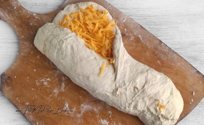 Place the flour, salt, sugar, and yeast in a bowl. Mix with a dry whisk. Add the oil and water. Stir with the back of a silicone spatula until the flour disappears. You will have a sticky dough. Cover the bowl with plastic wrap and let it rise for 1 1/2 hours in a warm, draft-free place. During this time, the dough should double in size.
Dust a pastry board with flour. Stretch the dough out onto the board. Place 2/3 of the cheese on the dough and spread it evenly over the surface.
Roll the dough into a roll along the short edge. Pinch the seam.